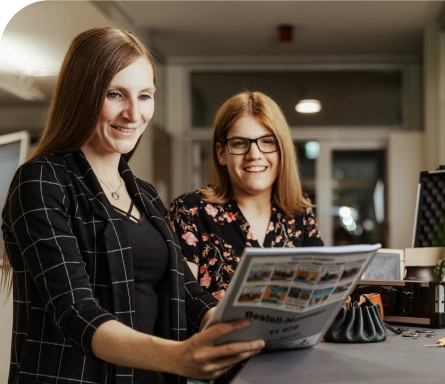 Zwei Kolleginnen lächeln und sehen sich gemeinsam ein Medienkatalog von dem KMZ Rottweil an.